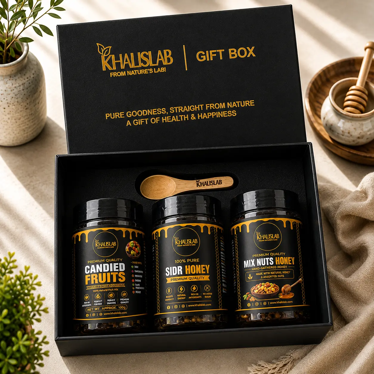 Khalislab gift box set containing three jars of honey and nuts: Candied Fruits, Sidr Honey, and Mix Nuts Honey, presented in an open black box with a wooden spoon, styled on a beige fabric surface with decorative plants and a honey dipper.