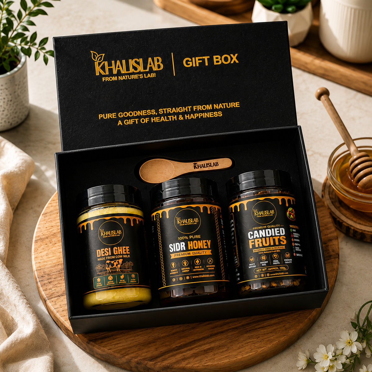 Khalislab gift box set containing three jars of ghee and honey, presented in an open black box on a wooden tray with a honey dipper and plants in the background.